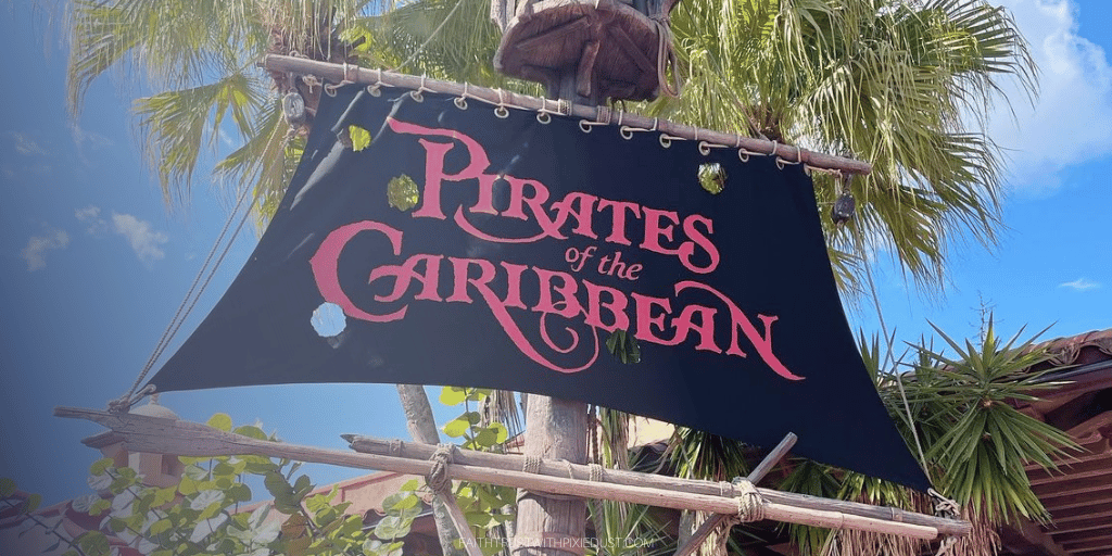 A weathered black banner with the text 'Pirates of the Caribbean' in ornate red lettering, hanging from a wooden frame with lush green leaves and palm trees in the background under a clear blue sky