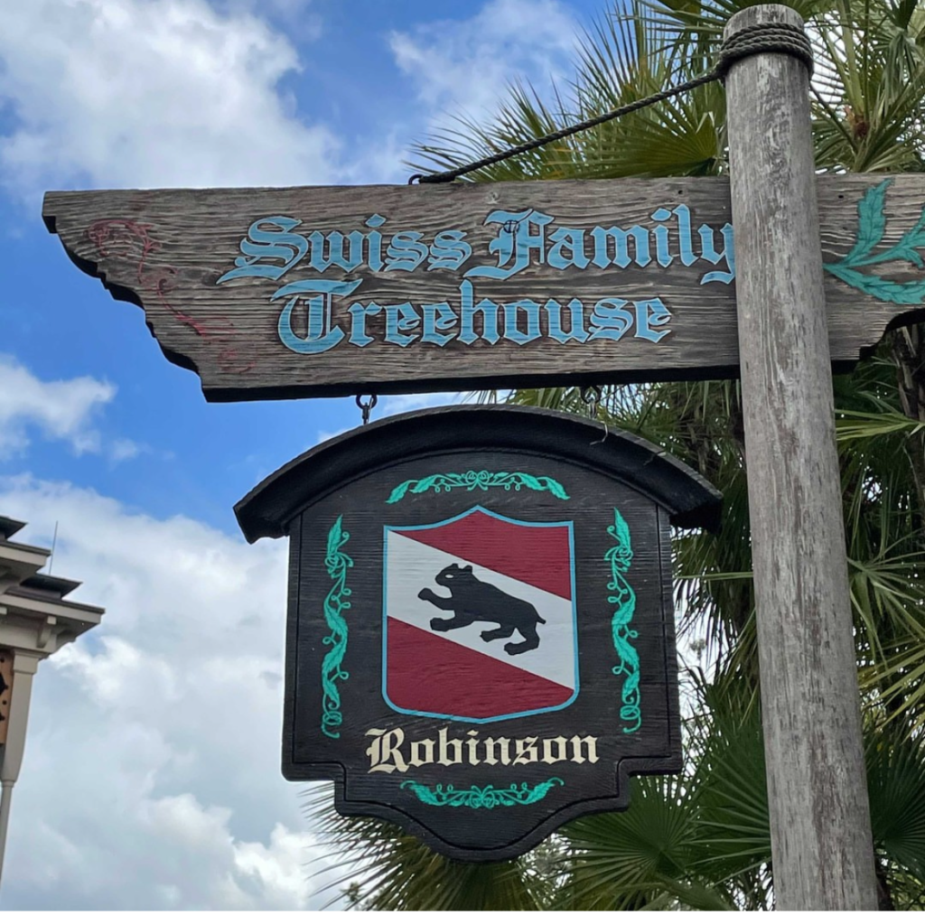 A rustic wooden sign reading 'Swiss Family Treehouse' with carved and painted leafy accents, suspended above a heraldic shield featuring a silhouette of a bear and the name 'Robinson,' set against a cloudy sky with palm fronds peeking into the frame."