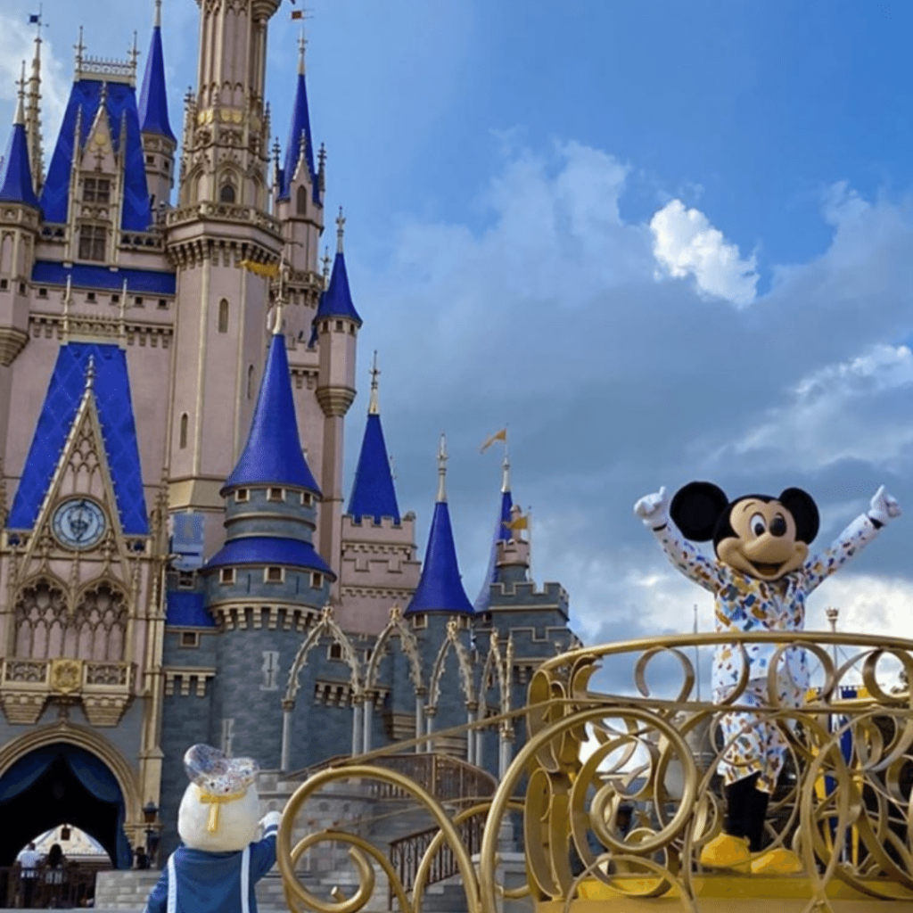 Mickey Mouse character waving happily in a white outfit with colorful decorations, standing on a golden float in front of the iconic Cinderella Castle with its distinctive blue rooftops and golden spires, under a partly cloudy sky at Disney