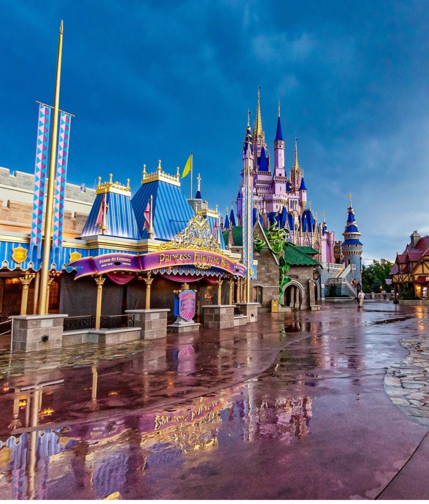 Cinderella Castle at Magic Kingdom gleams after a rain shower, with wet pavements reflecting its majestic spires and vibrant colors. The 'Princess Fairytale Hall' sign in the foreground adds a touch of enchantment to the scene under a brooding twilight sky.