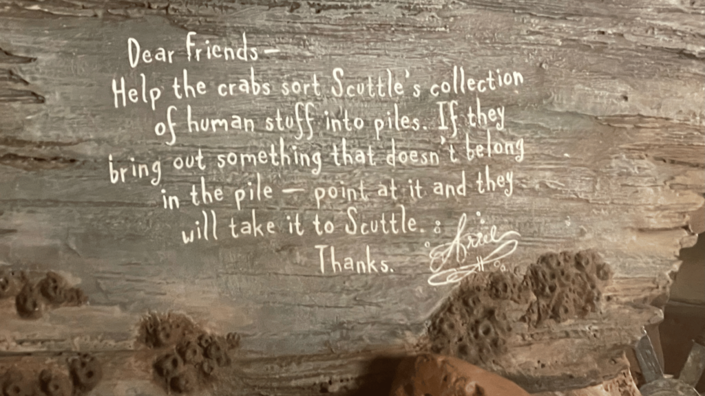A rustic, weathered wooden sign with a message for 'dear friends' asking for help to sort 'Scuttle's collection of human stuff into piles', with instructions to point at any out-of-place item so the crabs can take it to Scuttle, signed with thanks