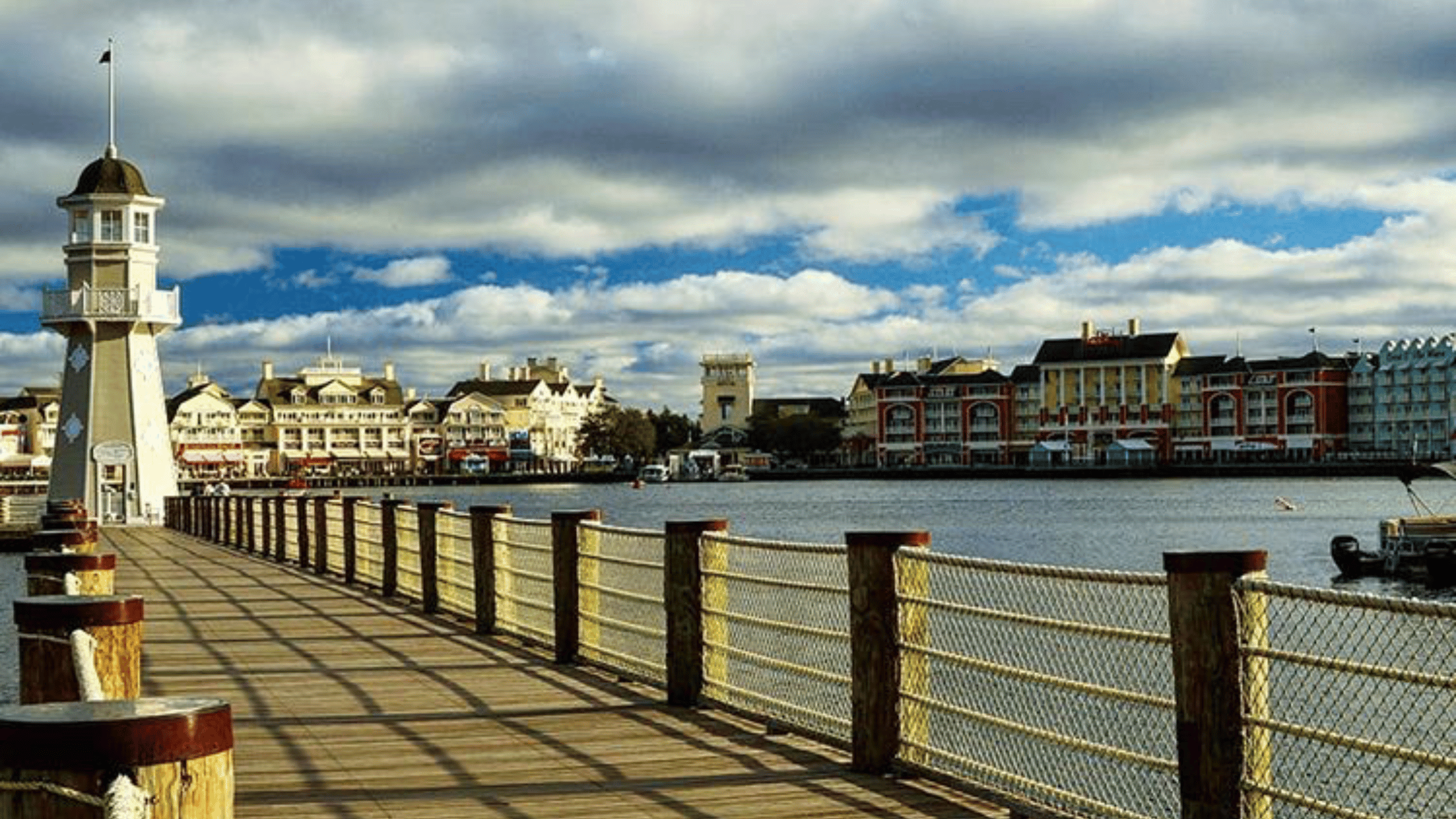 Disney boardwalk A scenic view of Disney's BoardWalk, featuring a lighthouse at the end of a wooden pier, with colorful resort buildings lining the opposite shore under a partly cloudy sky